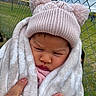 baby, child, blanket, hat, pom_poms, hand, outdoor, fence, grass, person, cold_weather, knitwear, face, expression, portrait, infant, warm_clothing, candid, closeup, nature
