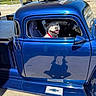 dog, vehicle, vintage_truck, blue_truck, pickup_truck, car_window, driver_seat, dog_face, smiling_dog, open_window, red_collar, outdoor, sunny, roadside, rural, blue_paint, chrome, reflection, classic_vehicle, automotive_photography