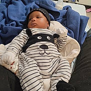 Jean Uriel a rejoint le concours — aidez-le/la à gagner de superbes lots ! baby, infant, bed, blanket, clothing, onesie, hat, mittens, bottle, furniture, interior, person, laying, striped_clothing, black_hat, white_mittens, baby_bottle, indoors, calm, young_child