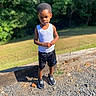 child, boy, toddler, outdoor, portrait, standing, forest, trees, grass, sunlight, summer, white_tank_top, black_shorts, sneakers, gravel, wood, nature, park, face, full_body