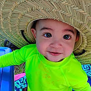 John is registered to the contest to win money with this photo: baby, child, infant, portrait, smiling, outdoor, sun_hat, straw_hat, green_shirt, bright_clothing, face, eyes, close_up, seated, blanket, blue_chair, summer, sunlight, playful, happy