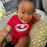 Zyair is registered to the contest to win money with this photo: baby, child, infant, smiling, red_onesie, curly_hair, pillow, couch, living_room, toe, hand, eyes, portrait, closeup, home_interior, television, sandal, blanket, happy, playtime