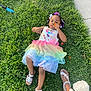 child, girl, grass, flower, colorful_dress, rainbow_tutu, sandals, outdoor, nature, greenery, playful, summer, cute, flowers, person, feet, relaxing, daylight, casual, happy
