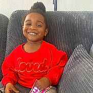 Zariah joined the competition — help win amazing prizes! child, toddler, smiling, red_sweater, indoor, couch, gray_cushion, lamp, holding_object, happy, portrait, casual_clothing, home, cozy, person, seated, hair_bun, bright_colors, young_child, card