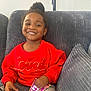 child, toddler, smiling, red_sweater, indoor, couch, gray_cushion, lamp, holding_object, happy, portrait, casual_clothing, home, cozy, person, seated, hair_bun, bright_colors, young_child, card