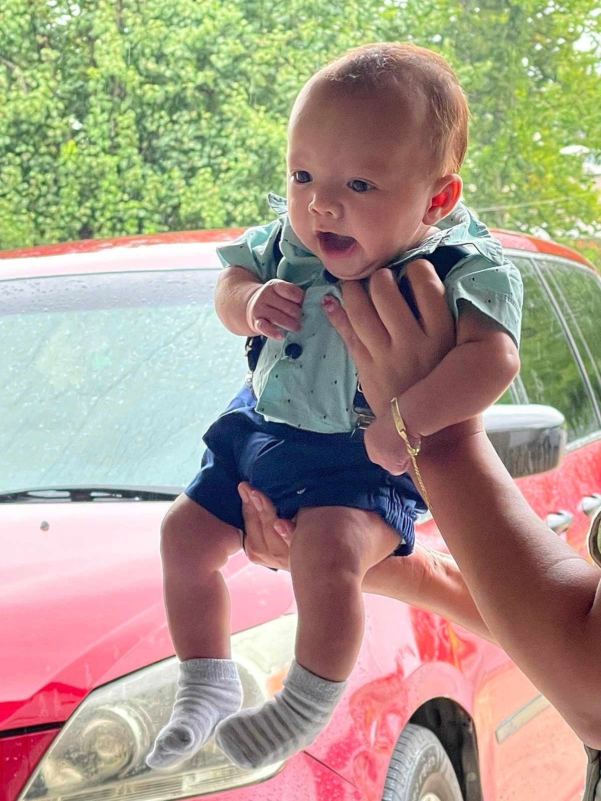 Elian Yandell Tejada is registered to the contest to win money with this photo: baby, child, person, hand, car, vehicle, red_car, greenery, outdoor, clothing, shorts, shirt, sock, holding, portrait, cute, happy, expression, human, skin
