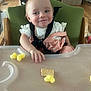 baby, high_chair, smile, child, food, snack, plastic_tray, clothing, indoor, person, wooden_floor, face, seat, straps, pink, white, hand, cute, happy, infant