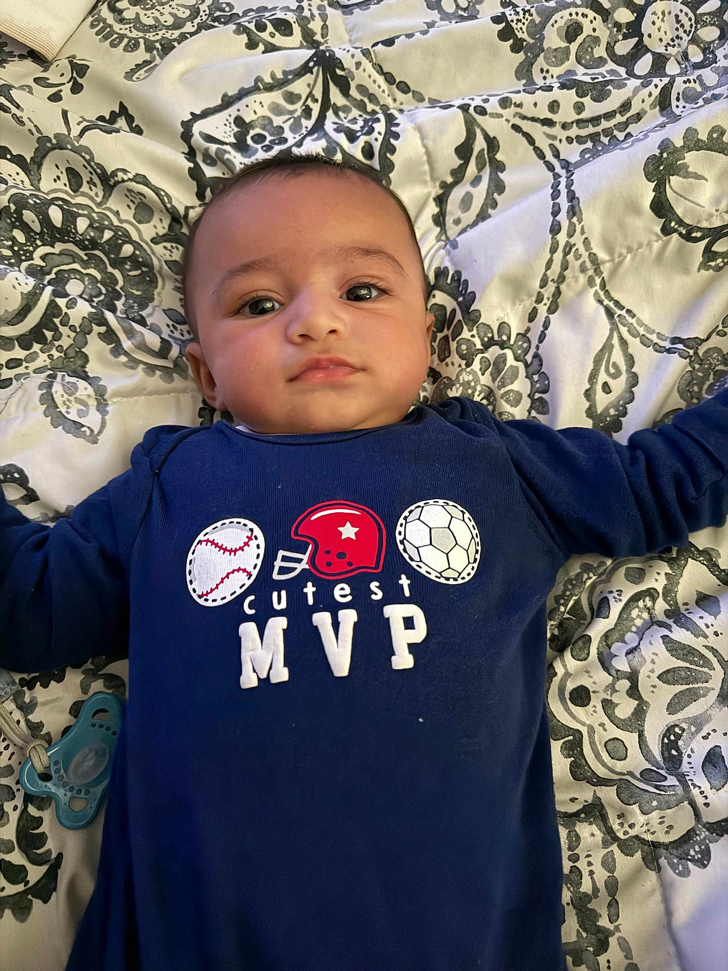 Saint joined the competition — help win amazing prizes! baby, infant, child, onesie, clothing, blue_clothing, face, head, lying_down, bedspread, pattern, pacifier, hand, arm, person, indoor, cute, young, portrait, expression