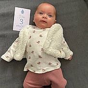 Maëlys participe au concours pour gagner de l'argent avec cette photo : baby, blanket, celebration, child, clothing, cute, fabric, face, indoor, infant, lying_down, milestone, milestone_card, pants, person, portrait, smile, socks, soft_texture, sweater