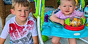 Riverlyn is registered to the contest to win money with this photo: child, baby, boy, smiling, happy, outdoor, baby_jumper, toy, wicker_chair, concrete_floor, fence, grass, playtime, casual_clothing, shorts, tshirt, barefoot, greenery, sunlight, family