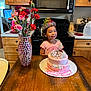 child, birthday, cake, tiara, flowers, vase, table, kitchen, oven, microwave, wooden_cabinet, smile, pink, party_hat, celebration, girl, happy, decor, indoor, person