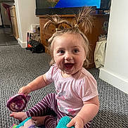 Celia joined the competition — help win amazing prizes! baby, carpet, child, cute, dimple, floor, hair_ties, happy, indoor, knitted_toy, living_room, person, pink_shirt, playful, pony_tail, smiling, striped_pants, television, toy, wooden_furniture