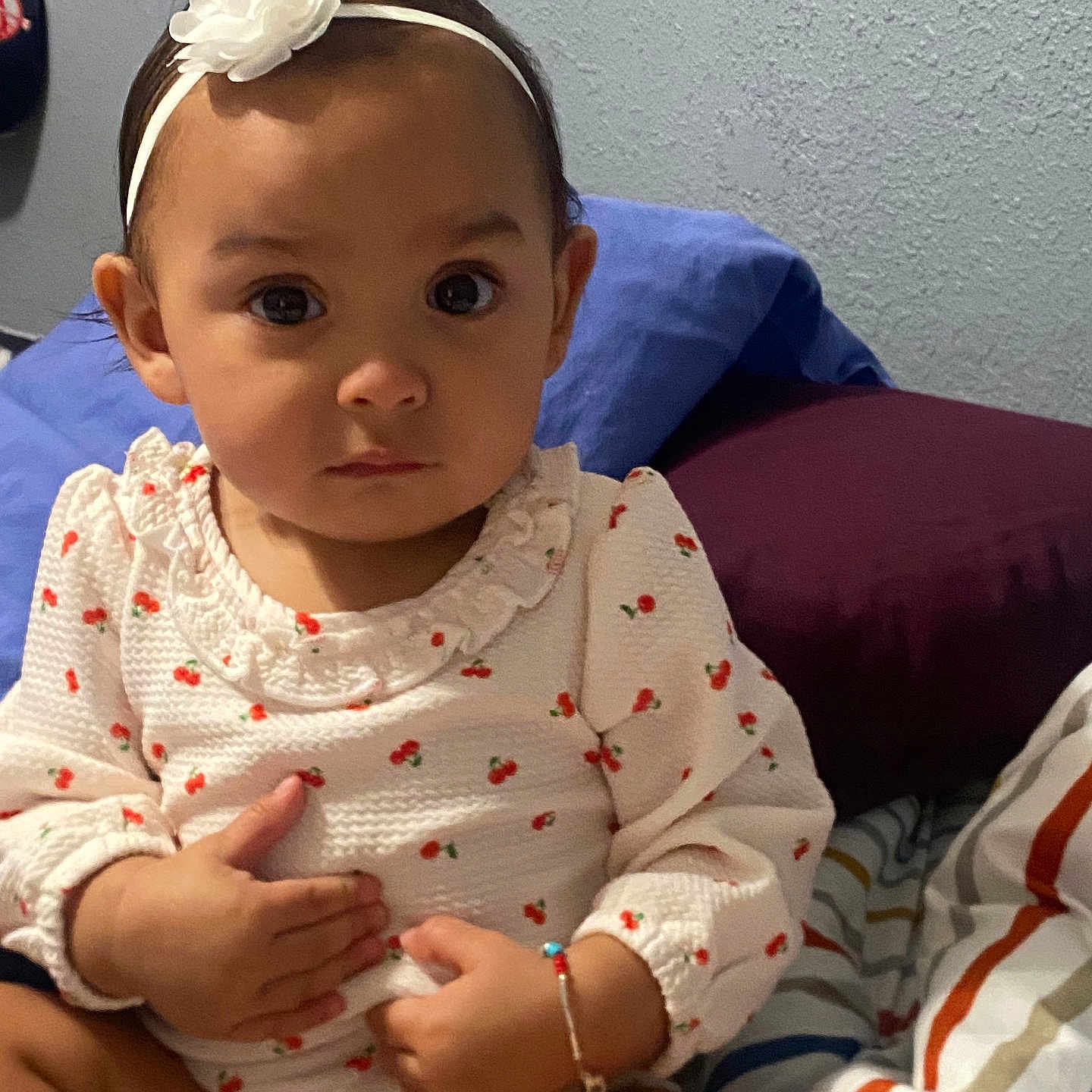 Aryana is registered to the contest to win money with this photo: baby, child, infant, portrait, sitting, bed, bedroom, headband, flower, dress, patterned_clothing, strawberries, wide_eyes, serious_expression, face, hands, bracelet, pillows, blanket, indoor