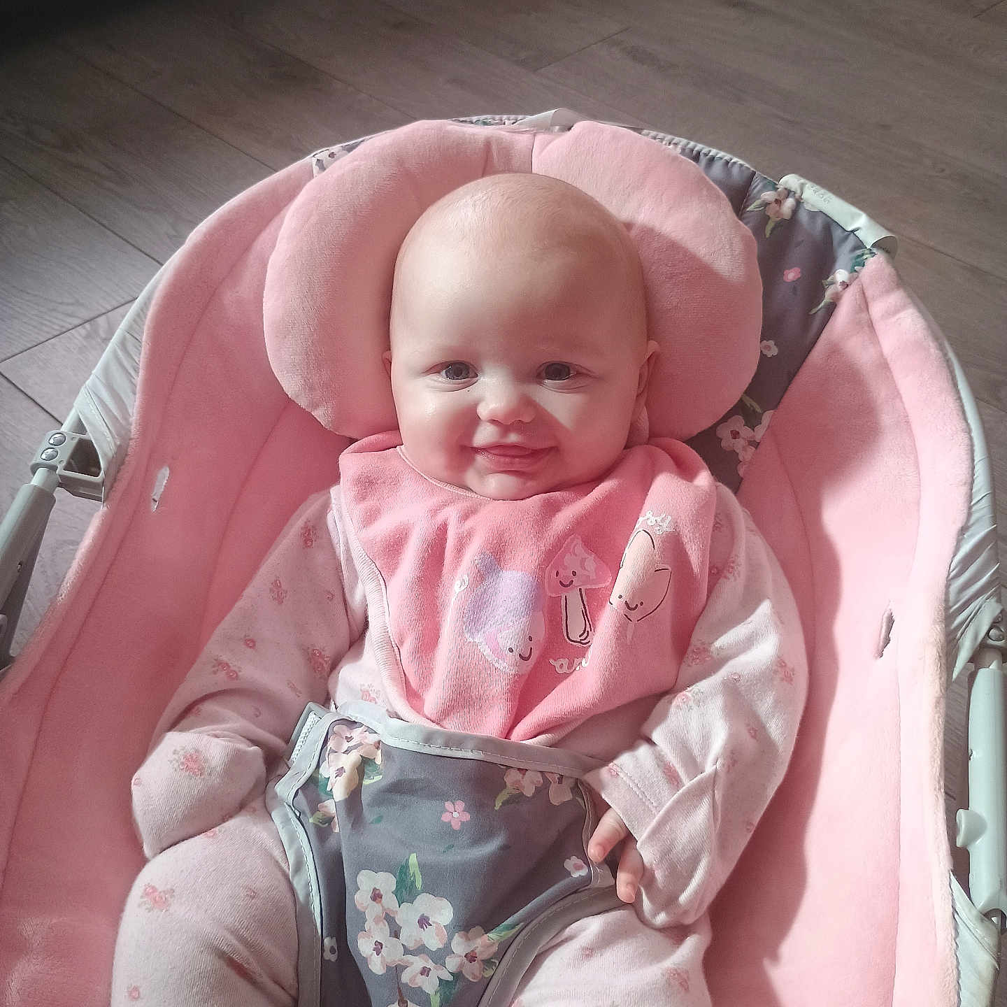 Ivy is registered to the contest to win money with this photo: baby, baby_chair, bib, child, comfort, cute, face, floral_pattern, happy, indoors, infant, pajamas, person, pink, plush, portrait, seated, smiling, soft_toys, wooden_floor