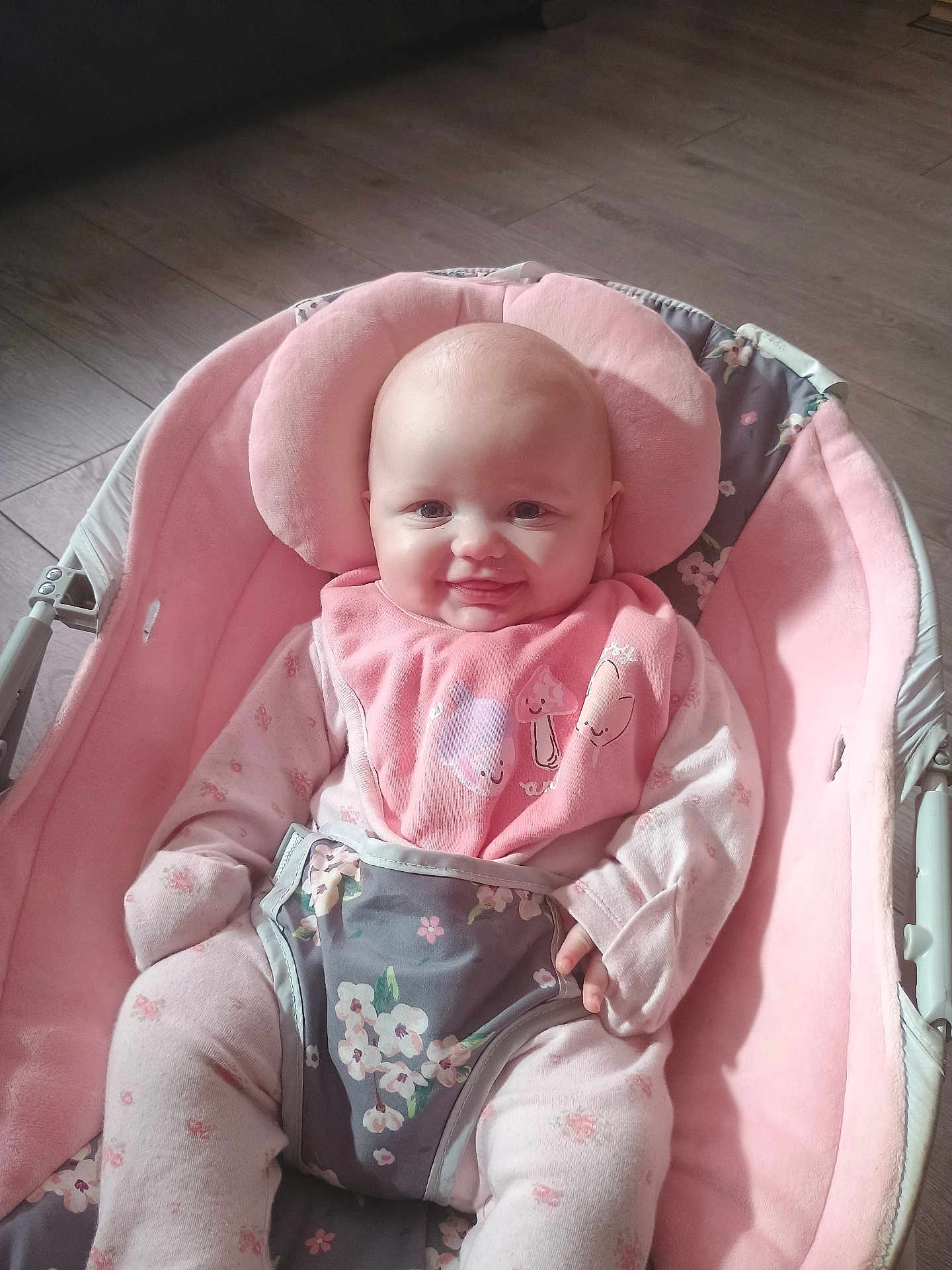 Ivy is registered to the contest to win money with this photo: baby, infant, smiling, pink, bib, baby_chair, pajamas, floral_pattern, face, person, seated, indoors, wooden_floor, cute, child, soft_toys, plush, comfort, happy, portrait