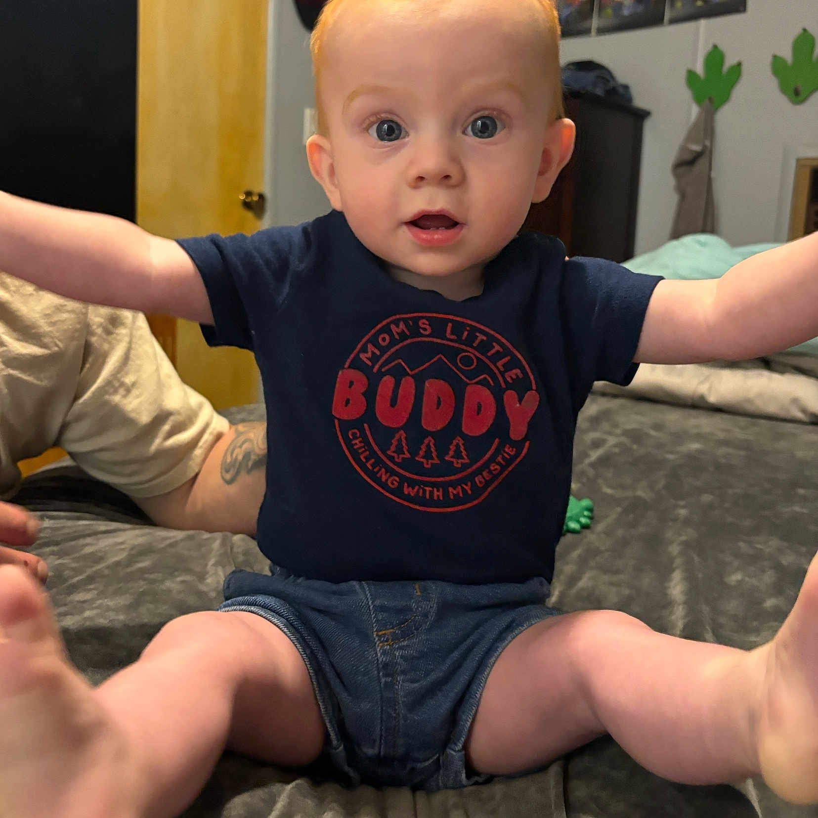Dino is registered to the contest to win money with this photo: arms, baby, bed, bedroom, child, clothing, denim_shorts, door, expression, eyes, face, furniture, indoor, infant, legs, mouth, navy_shirt, person, tattoo, text_on_clothing