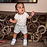 Aiden is registered to the contest to win money with this photo: toddler, child, person, white_tshirt, gray_shorts, sneakers, gold_chain, bracelet, couch, cushions, patterned_cushions, mirror, indoor, standing, curious_expression, brown_couch, footwear, shorts, jewelry, home