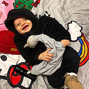 Noah is registered to the contest to win money with this photo: baby, child, smiling, hoodie, black_clothing, toy, bedspread, colorful, moccasins, footwear, blanket, indoor, happy, face, person, clothing, hand, fabric, lying_down, cute