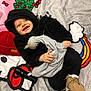 baby, child, smiling, hoodie, black_clothing, toy, bedspread, colorful, moccasins, footwear, blanket, indoor, happy, face, person, clothing, hand, fabric, lying_down, cute
