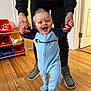 baby, child, person, indoor, wooden_floor, toy_bins, smiling, holding_hands, blue_onesie, curly_hair, happy, walking, adult_hands, footwear, door, flooring, clothing, home, playroom, cute