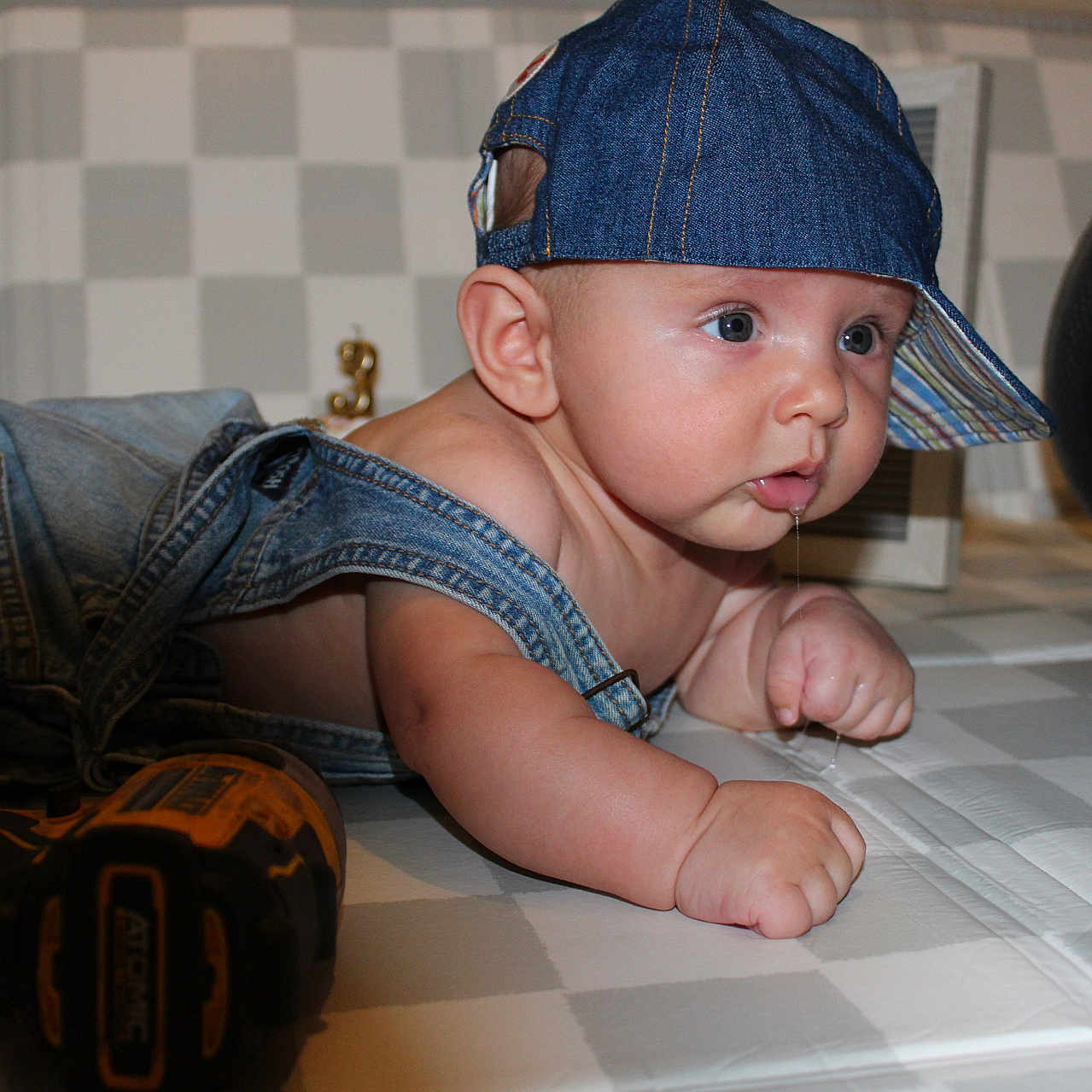 Croix is registered to the contest to win money with this photo: baby, denim, cap, overalls, drool, tools, wrench, drill, measuring_tape, checkered_floor, curious, infant, face, hand, floor, indoor, person, child, blue, hat