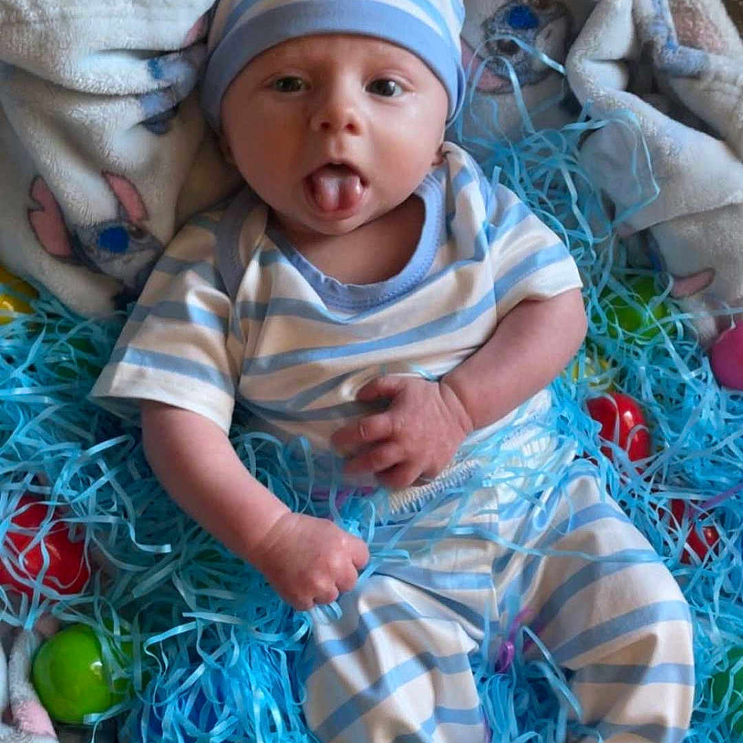 James joined the competition — help win amazing prizes! baby, infant, striped_clothing, hat, blanket, blue, white, plastic_eggs, shredded_paper, playful, tongue_out, hand, feet, cozy, cute, lying_down, indoors, colorful, soft_texture, smiling