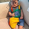 Koda joined the competition — help win amazing prizes! accessory, baby, bare_feet, child, colorful_clothing, couch, cute, dollar_pendant, expressionless, glasses, gold_chain, indoors, infant, living_room, overalls, playful, portrait, sitting, sofa, striped_shirt