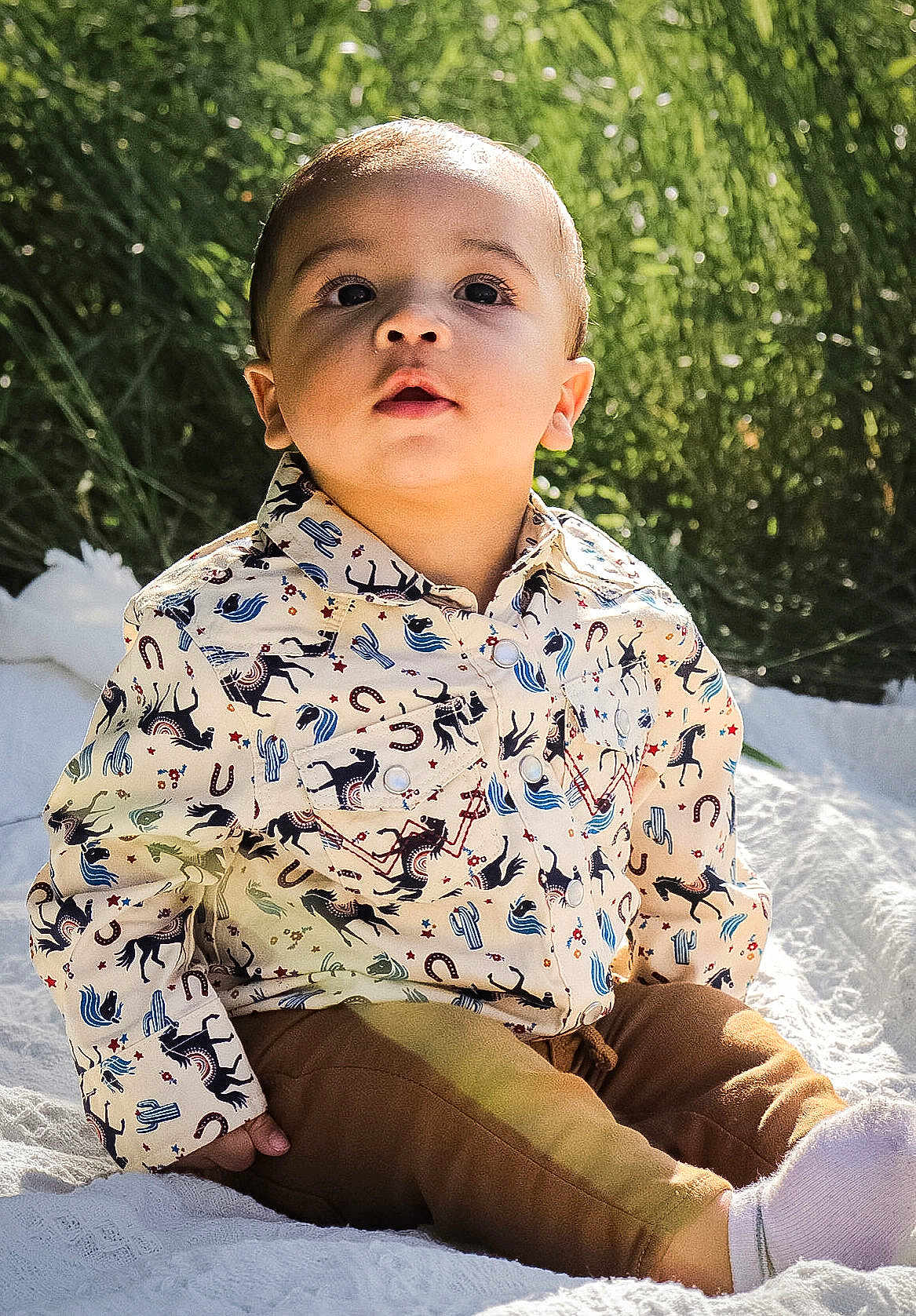 Kolson is registered to the contest to win money with this photo: toddler, child, outdoor, grass, blanket, shirt, patterned_clothing, brown_pants, socks, sitting, sunlight, face, curious, young_child, portrait, nature, baby, cute, daylight, expression