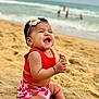 baby, beach, sand, ocean, water, waves, smiling, child, headband, red_clothing, summer, vacation, happy, sitting, cute, outdoor, nature, sunny, playful, toddler