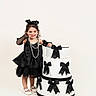 child, girl, black_dress, pearl_necklace, black_bow, decorative_cake, white_cake, gloves, white_shoes, studio_shot, playful_pose, smiling, cute, birthday_theme, fashion, portrait, indoor, celebration, young_child, accessories