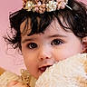 baby, child, face, portrait, curly_hair, flower_crown, blanket, pink_background, cute, close_up, innocent, soft_texture, adorable, young_child, smiling, headband, skin, person, toddler, warmth