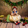 baby, child, apple, costume, dress, headband, smile, toy, mushroom, plush, floor, greenery, decor, portrait, sitting, red, blue, wood, cushion, indoor