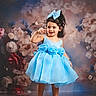 child, girl, blue_dress, bow, curly_hair, floral_background, flowers, posing, smiling, portrait, fashion, dress, footwear, standing, studio_photo, cute, happy, accessories, young_child, indoor