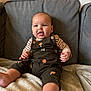 Theodore is registered to the contest to win money with this photo: baby, child, overalls, mushroom_pattern, striped_shirt, couch, blanket, indoor, smiling, sitting, face, feet, hand, comfortable, soft_texture, young_child, portrait, cozy, home, infant