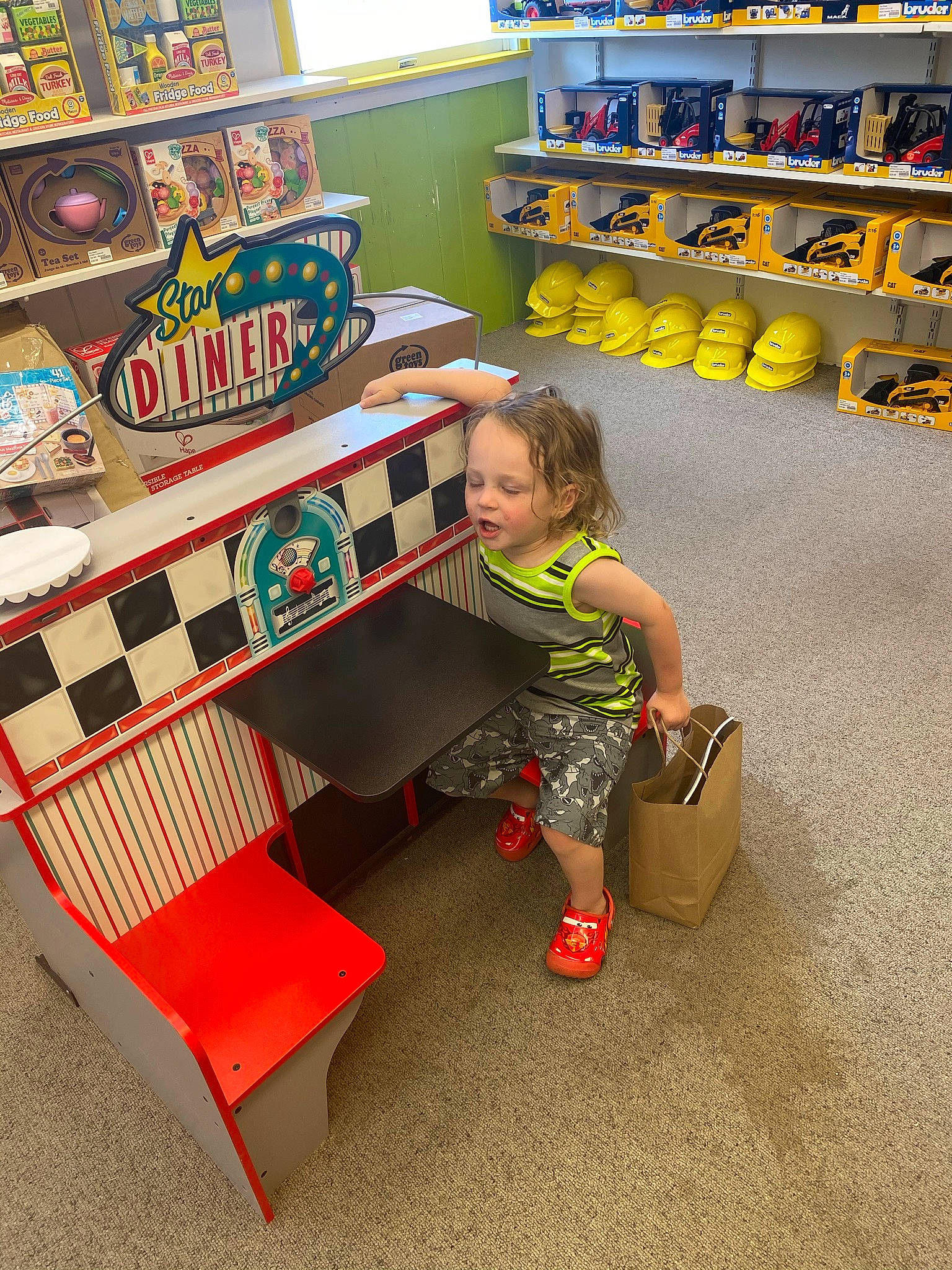 Jd is registered to the contest to win money with this photo: bench, bookcase, child, convenience_store, couch, flooring, fun, person, play, retail, room, shelf, shelving, sitting, t_shirt, table, tire, toddler, toy, toy_vehicle