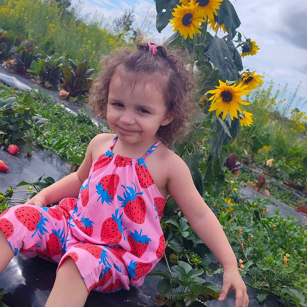 Velenia is registered to the contest to win money with this photo: casual_clothing, child, curly_hair, daylight, field, flowers, garden, girl, greenery, growth, nature, outdoor, plants, playful, smiling, sneakers, strawberry_print, summer, sunflowers, vegetation