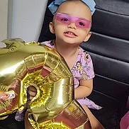 Velenia is registered to the contest to win money with this photo: balloon, birthday, black_chair, blue_bow, casual_clothing, celebration, child, fun, gold_balloon, happy, holding, indoors, number_three, party, person, pink_sunglasses, playful, seated, smiling, young_child