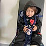 toddler, child, hoodie, black_clothing, sitting, baby_seat, straps, indoor, chair, floor, curious, expression, casual, footwear, white_socks, patterned_fabric, young_child, portrait, person, home