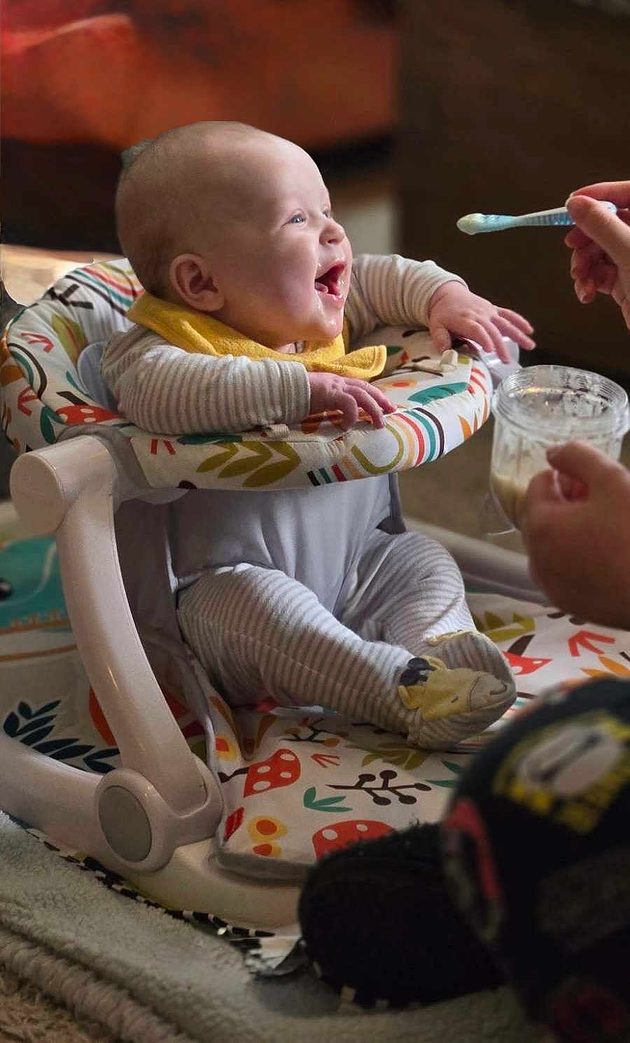 Ezra joined the competition — help win amazing prizes! baby, bib, closeup, cute, eye_contact, feeding, food, hands, highchair, home_interior, infant, jar, laughing, onesie, parent_hand, patterned_fabric, play_seat, smile, socks, spoon