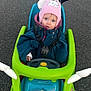 baby_clothing, child, cold_weather, cute, green_frame, looking_up, outdoors, pavement, person, pink_hat, portrait, ride_on_toy, safety_harness, seat, shoes, steering_wheel, teddy_bear, toddler, toy_car, winter_coat