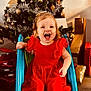 bauble, child, christmas_tree, festive, gift, holiday, indoor, laughing, ornament, portrait, present, red_dress, shoes, slide, smile, tinsel, toddler, toy, wooden_floor, wrapped_gift