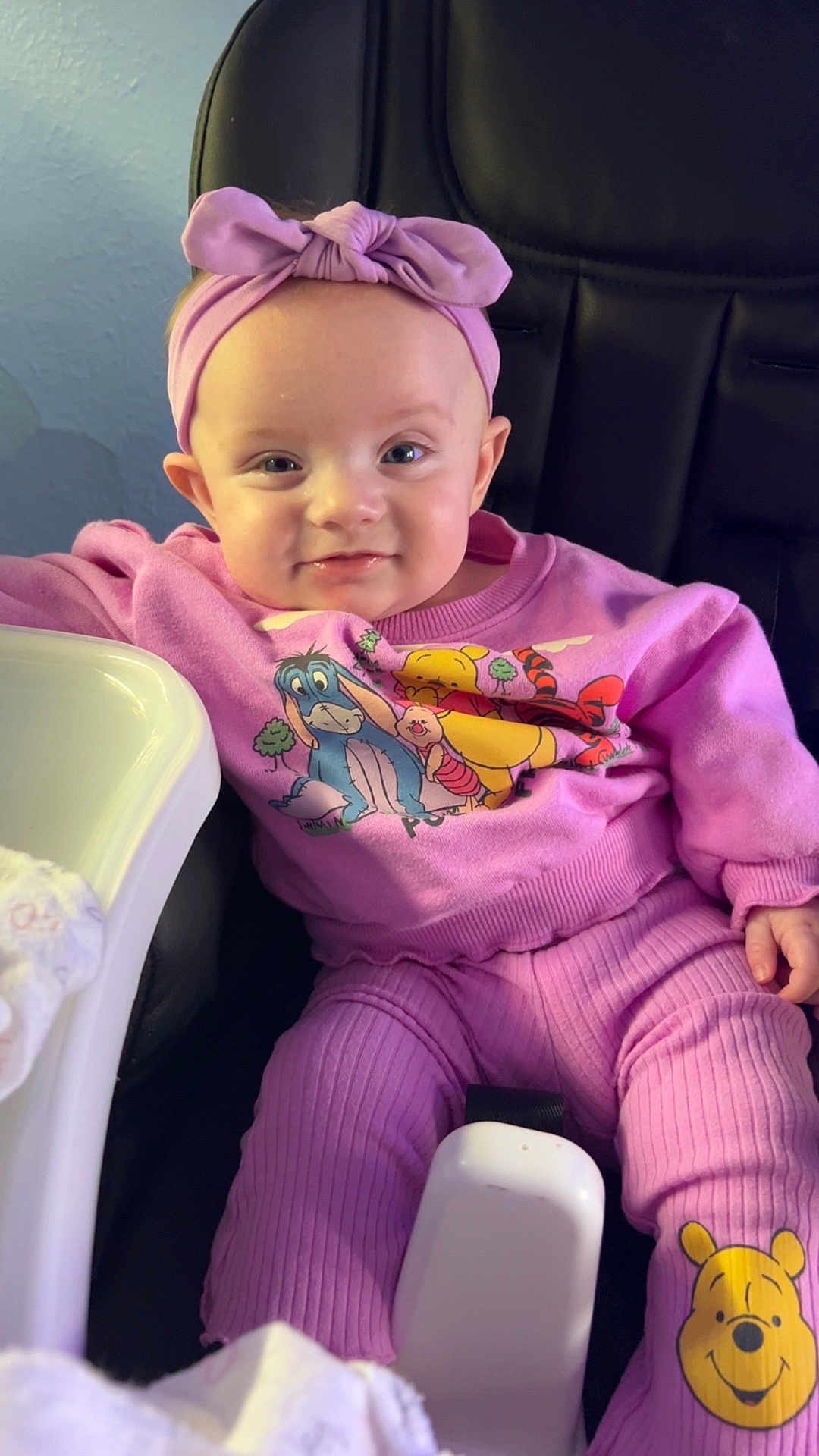 Izabel is registered to the contest to win money with this photo: baby, child, purple_outfit, headband, winnie_the_pooh, high_chair, smiling, infant, cute, seated, indoors, clothing, person, face, apparel, toddler, expression, comfort, soft_lighting, playful