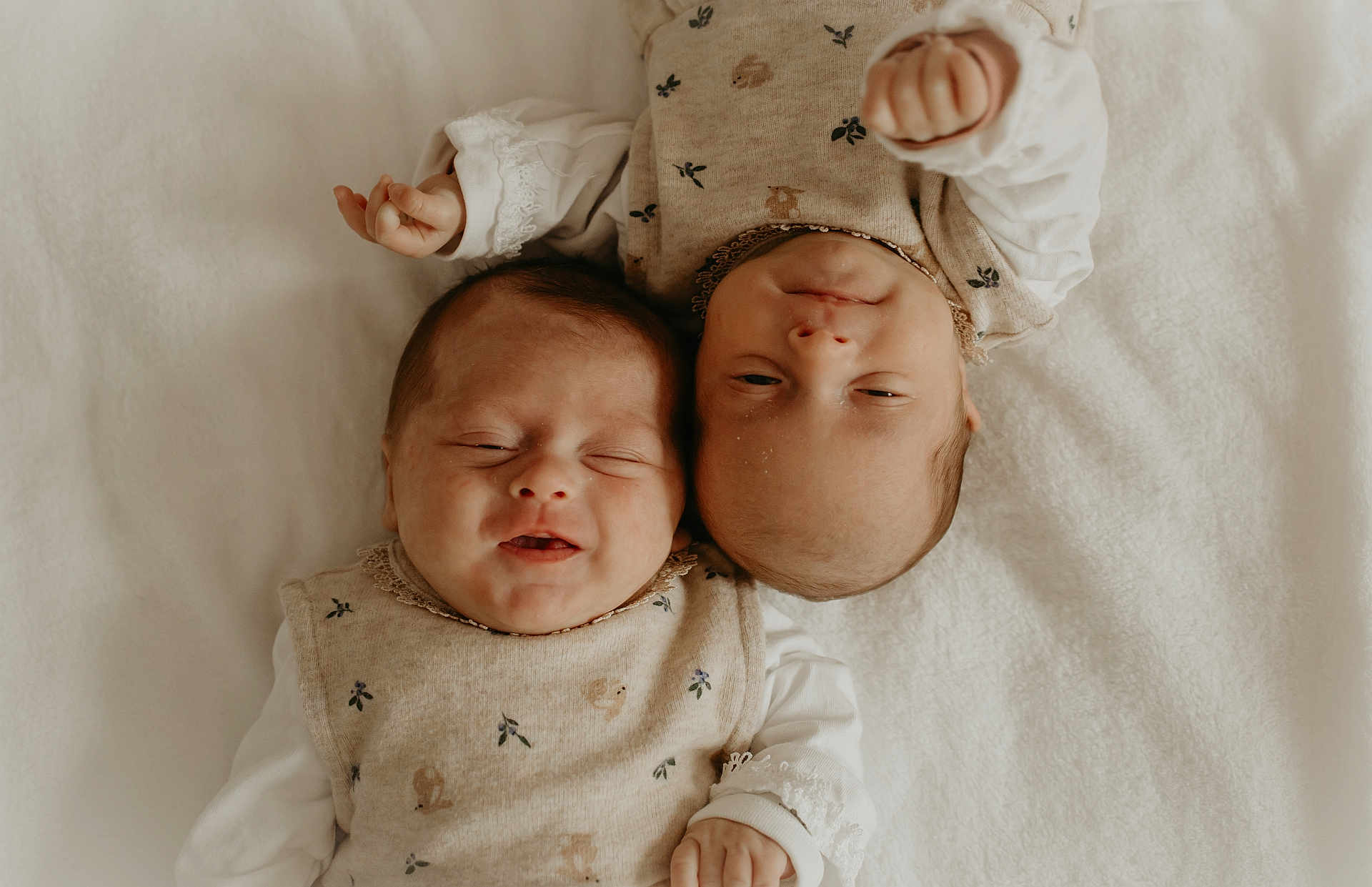 Louise Et Lola participe au concours pour gagner de l'argent avec cette photo : baby, infant, newborn, two_babies, lying_down, white_blanket, matching_outfits, beige_clothing, floral_pattern, smiling, peaceful, cute, sleepy, portrait, soft_lighting, indoor, closeup, child, head, hands
