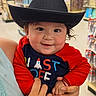 baby, child, cowboy_hat, smile, red_clothing, onesie, person, indoor, store, aisle, hand, holding, cute, face, young_child, toy_store, colorful, clothing, happy, portrait