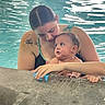baby, woman, swimming_pool, water, tattoo, toy, child, person, swimsuit, playing, holding, curious, interaction, family, poolside, skin, wet, summer, fun, bonding
