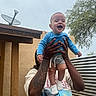 baby, child, person, hands, tattoo, shirt, blue_clothing, smiling, outdoor, tree, sky, satellite_dish, building, wood, metal, fence, happy, expression, holding, portrait