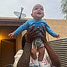 Andre is registered to the contest to win money with this photo: baby, child, person, hands, tattoo, blue_shirt, shorts, socks, outdoor, building, door, satellite_dish, metal_wall, sky, smiling, lifting, happy, arm, portrait, casual