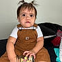 baby, bed, blanket, brown_clothing, child, colorful_clips, expression, face, hair_ties, hands, indoor, jar, overalls, person, pigtails, pillow, sitting, toddler, white_shirt, young_child