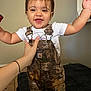 toddler, child, smiling, happy, overalls, camo, white_tshirt, indoor, person, baby_teeth, hand, supporting, arms_up, face, hair, floor_tiles, blanket, couch, wall, home