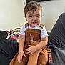 baby, blanket, brown_clothing, casual, child, cute, face, furniture, hand, happy, holding, indoor, overalls, person, portrait, sitting, smile, television, toddler, white_shirt